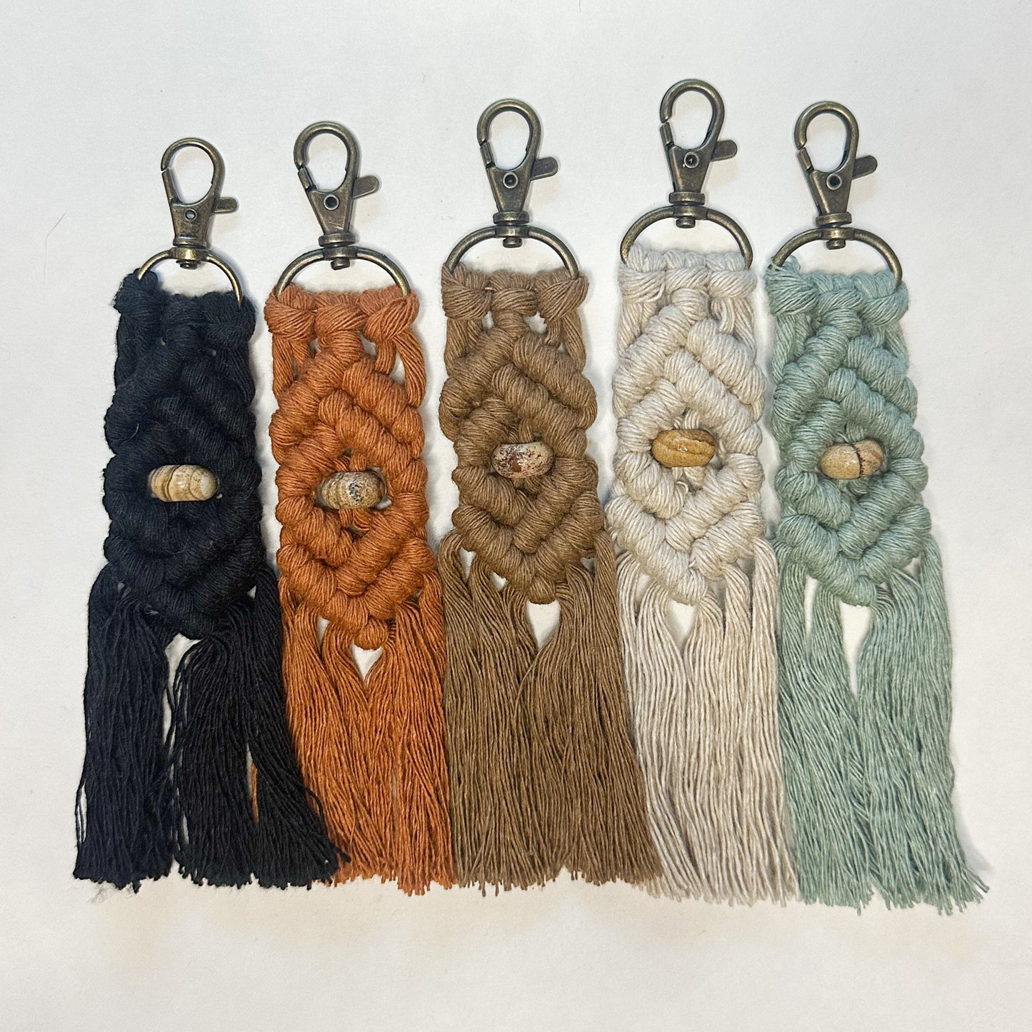 Five macrame keychains in black, brown, beige, white, and green on a light gray background.