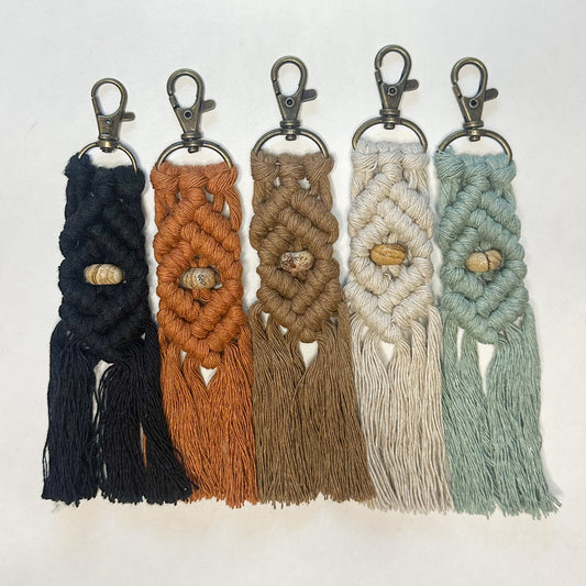 Five macrame keychains in black, brown, beige, white, and green on a light gray background.
