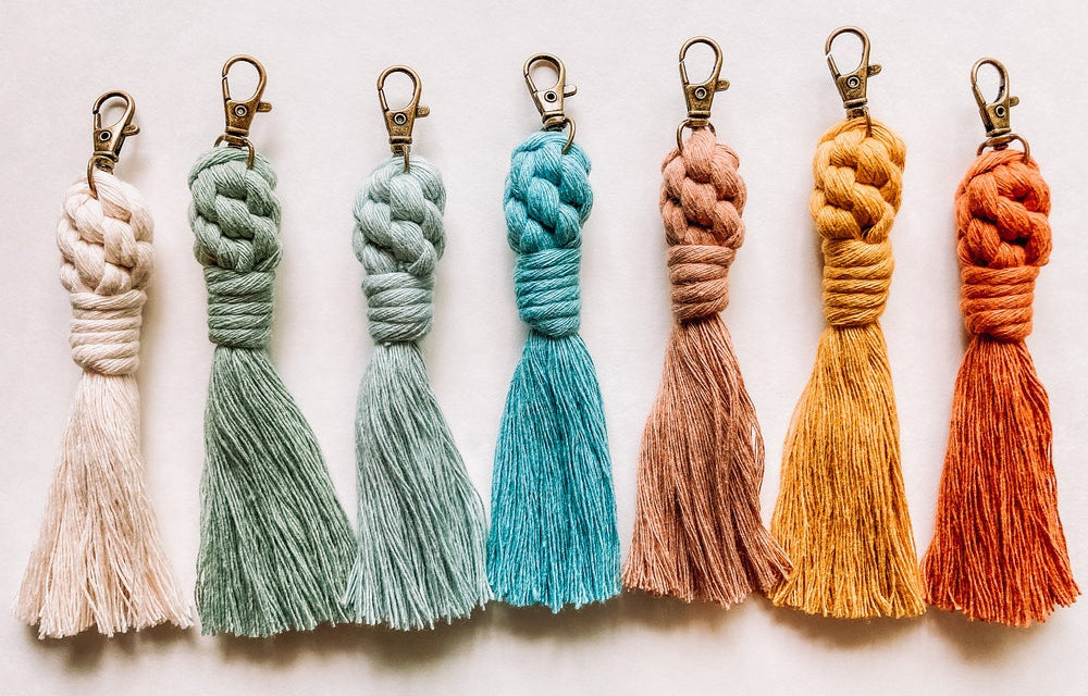 A collection of handcrafted macrame tassel keychains in various colors, including beige, blue, gray, orange, pink, and brown, displayed on a white background.